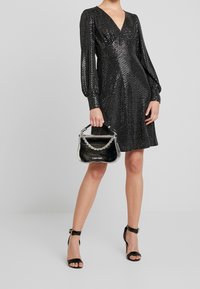 Černé flitrové šaty s dlouhými nabíranými rukávy, hlubokým V-výstřihem a přiléhavým pasem, doplněné malou černou lakovanou kabelkou s řetízkovým popruhem.