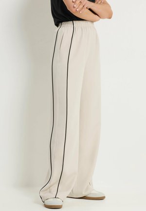 Personne portant un pantalon beige ample avec des bandes latérales noires et des chaussures blanches, les bras croisés, sur un fond clair uni.