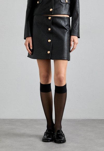 Black leather mini skirt with gold button detailing, paired with black knee-high socks and loafers, showcasing a sleek, structured design.