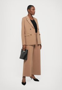 Beige dobbeltradet blazer med gullknapper, kombinert med matchende brede bukser. En svart håndveske og spisse svarte hæler fullfører antrekket.