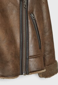 Section d'une veste en cuir marron avec fermeture éclair centrale argentée, poche zippée inclinée, bordure noire et revers en peau lainée marron clair visibles.