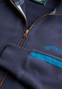 Sudadera azul marino con cremallera, con textura acanalada, cremallera dorada, acento de bolsillo en azul claro y "SUPERDRY" bordado en azul.