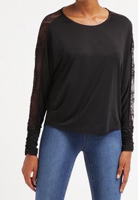 Czarny, długi rękaw bluzka z koronkowymi akcentami na ramionach, okrągłym dekoltem i luźnym kroju. W zestawie z niebieskimi dżinsami. Gładka tekstura.