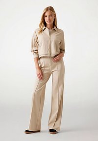 Veste beige courte zippée avec deux poches avant, associée à un pantalon taille haute à jambes larges, tous deux en tissu lisse. Chaussures plates noires.