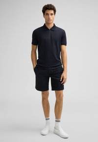 Granatowa koszulka polo z zamkiem przy kołnierzyku, połączona z teksturowanymi granatowymi szortami. Uzupełniona o białe buty wsuwane i paskowane skarpetki do kostek.