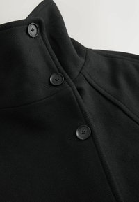 Geacă neagră cu guler înalt, realizată dintr-un material texturat, cu trei nasturi mari negri și cusături ajustate pentru un design structurat.