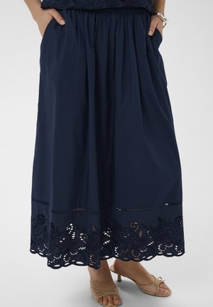 Femme portant une jupe midi bleu marine avec un détail floral découpé à l'ourlet et des sandales à talons bas ouvertes de couleur beige, les mains dans les poches.