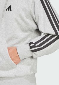 Šedá mikina s kapucňou má čierne tri pruhy na rukávoch a logo. Mäkký materiál s predným vreckom a rebrovanými manžetami.