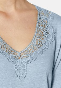 Lichtblauwe blouse met een gedetailleerde kanten halslijn met ingewikkelde bloemenpatronen en gegolfde randen, gemaakt van zachte, gestructureerde stof.