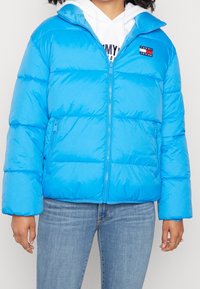 Bright blue puffer jacket with a zip front, high collar, and stitched paneling. Features a logo patch and side zip pockets.