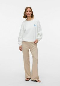 Sudadera blanca con bordado azul de "Tuscany", combinada con pantalones de pierna ancha en color beige. La sudadera tiene hombros caídos y un corte relajado.