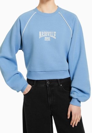 Light blue long-sleeve sweatshirt with "Nashville 1991" text, paired with black high-waist pants, one hand in pocket and one at side.
