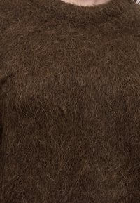 Gros plan d’une personne portant un pull en maille épaisse, marron, duveteux, avec un col rond.