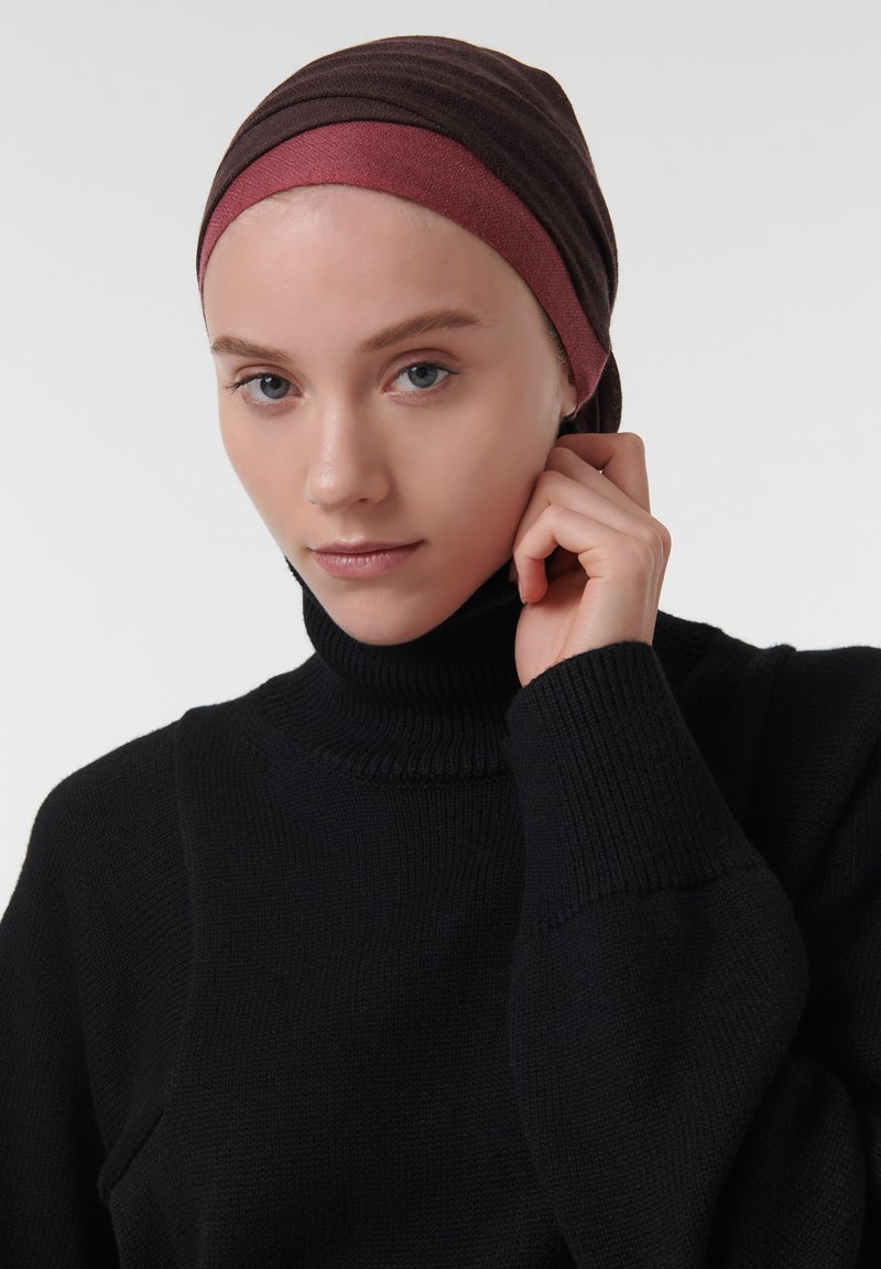 Pañuelo de cabeza marrón oscuro y burdeos con tejido texturizado, usado con un suéter de cuello alto negro con relieve. Primer plano de la cara, manos ajustando el pañuelo.