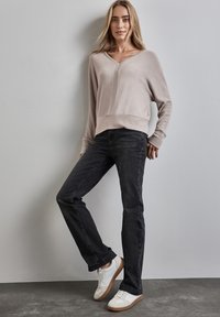 Beiger, übergroßer Pullover mit V-Ausschnitt und gerippten Bündchen, kombiniert mit dunklen Jeans und weißen Turnschuhen mit einer beigefarbenen Sohle. Schlichtes und lässiges Design.
