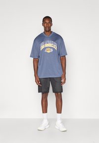 Blå mesh t-shirt med kort ärm och texten "Los Angeles Lakers"; ihop med svarta shorts och vita träningsskor.