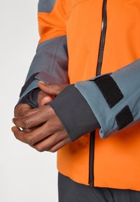 Τα χέρια είναι σταυρωμένα μπροστά, φορώντας ένα φωτεινό πορτοκαλί μπουφάν με γκρι μανίκια και μαύρες ρυθμιζόμενες αυτοκόλλητες ζώνες στους καρπούς.