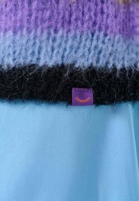 Pletený sveter v purpurovej, modrej a čiernej farbe s jemnou textúrou. Obsahuje prišitú purpurovú etiketu s oranžovým slnkom na leme.