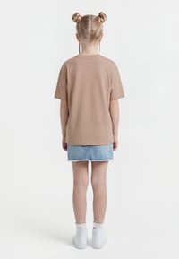 Otrok s svetlimi lasmi v dveh figah, oblečen v bež majico, denim krilo, bele nogavice in bele superge, stoji obrnjeno stran.