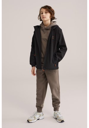 Kind staand in een bruine hoodie en joggingbroek, met een zwarte ritssluiting jas en witte sneakers tegen een effen achtergrond.