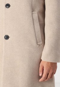 Beige wool coat with a soft texture, featuring black buttons and a side pocket. The design is tailored with a straight silhouette.