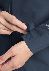 Giacca impermeabile navy con superficie liscia, dotata di polsini regolabili e un tirante in gomma per una vestibilità sicura.