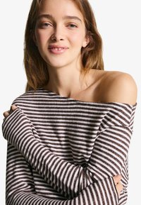 Prugasti pulover s dizajnom bez rukava, izrađen od mekog materijala. Ima horizontalne smeđe i bijele pruge, duge rukave s uskim manžetama.