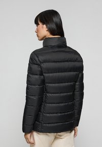 Giacca a vento nera con colletto alto, caratterizzata da sezioni trapuntate e una superficie liscia e lucida. Silhouette aderente, con dettagli minimi.