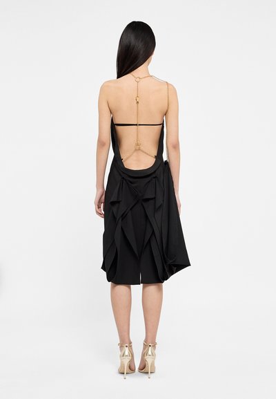 Črna obleka z globokim izrezom na hrbtu in zlate verige, asimetrične spodnje robove ter večplastni naborki. Gladka tekstura in prilega telo.