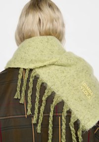 Persona rubia con el cabello corto, que lleva un abrigo marrón de cuadros y una bufanda verde claro con flecos y una etiqueta con la marca "BIMBA Y LOLA".