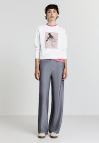 Felpa bianca con una grafica rosa, sovrapposta a una maglietta rosa. Pantaloni grigi a gamba larga con un motivo strutturato. Sneakers bianche.