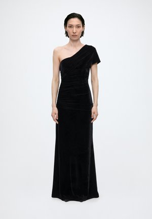 Femme debout portant une longue robe de soirée en velours noir à une épaule avec des détails froncés sur un fond blanc uni.