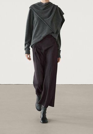 Osoba spacerująca w ciemnoszarej, luźnej bluzie, szerokich czarnych spodniach i czarnych botkach na gładkiej beżowej podłodze na tle białej ściany.