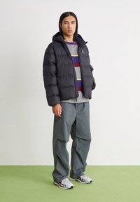 Svart puffjacka med huva, lättviktsmaterial, quiltat mönster. Kombinerad med grå randig tröja och grå byxor. Sneakers med accentfärger.