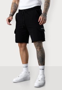 Shorts cargo noirs avec un ourlet effiloché, une taille élastique et des poches latérales. Associés à des baskets blanches et des chaussettes texturées.