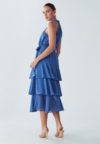 Plava haljina s više slojeva koja ima bezrukav dizajn, pojasan struk i lepršave slojeve. Tkanina je lagana i lagano se kreće.