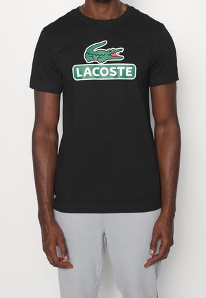 Mężczyzna w czarnej koszulce z zielono-białym logo Lacoste i szarych spodniach stoi na jednolitym tle.
