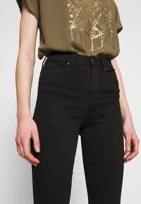 Jeans negros de cintura alta, hechos de mezclilla. Presentan un estilo clásico de cinco bolsillos y un acabado liso. Combinados con una blusa texturizada en color oliva.