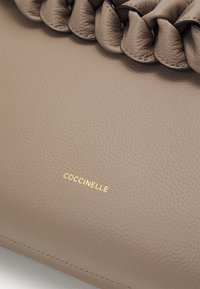 Beige Coccinelle-Handtasche, strukturiertes Leder, mit geflochtenem Griff und goldfarbenem Logo. Abgerundete rechteckige Form mit sauberen Stickdetails.