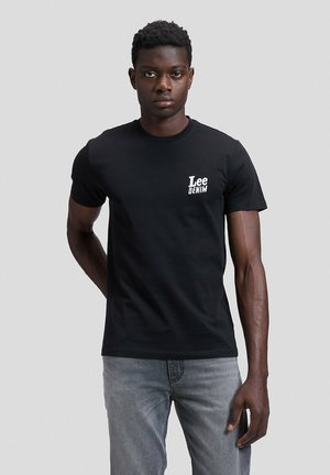 Hombre de pie frente a un fondo liso, lleva una camiseta negra Lee Denim y vaqueros gris claro, mirando al frente.