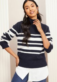 Navy- und weiße gestreifte Sweater mit hohem Kragen, gerippten Bündchen und Saum, über einem weißen Hemd getragen. Weiche Textur und lässiges Design.
