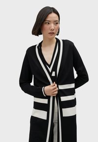 Svart tröja med långa ärmar, v-ringning, med vita geometriska mönster och ränder. Tyget verkar slätt med en avslappnad passform.