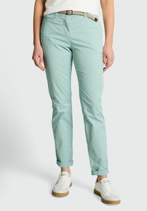 Person trägt hellblaue hochgekrempelte Hose, beigen gewebten Gürtel, weiße Sneakers und ein in die Hose gestecktes weißes Oberteil vor einfarbigem Hintergrund.