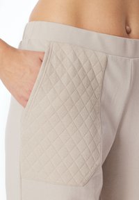 Béžové kalhoty s texturovanou, prošívanou kapsou na levé straně. Materiál se zdá být měkký a hladký, což zvyšuje celkový design.