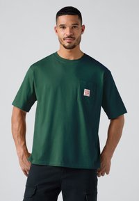 Mand med kort hår og skæg iført mørkegrøn T-shirt med korte ærmer og brystlomme samt sorte bukser, står imod en ensfarvet lys baggrund.