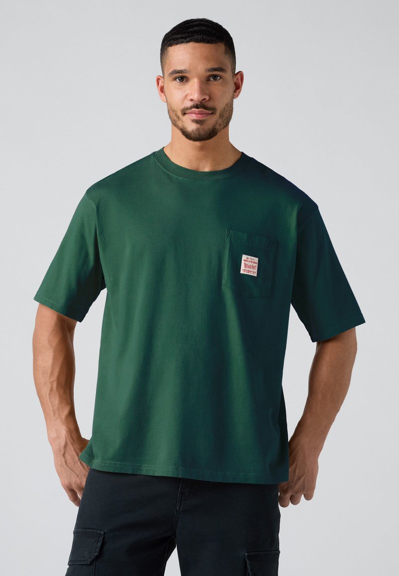Mand med kort hår og skæg iført mørkegrøn T-shirt med korte ærmer og brystlomme samt sorte bukser, står imod en ensfarvet lys baggrund.