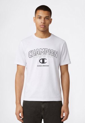 Camiseta blanca de algodón con un gráfico llamativo "CHAMPION" en negro y gris con un pequeño logotipo debajo. Mangas cortas y cuello redondo.