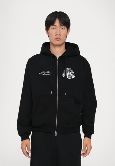Junger Mann trägt einen schwarzen Reißverschluss-Hoodie mit weißen Trompetengrafiken und dem Text "Filling Pieces United By Rhythm" auf der Brust und steht still.