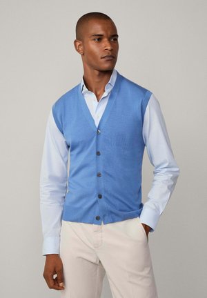 Gilet a maglia azzurro chiaro con scollo a V, dotato di sei bottoni scuri sul davanti, indossato sopra una camicia azzurro chiaro con bottoni e abbinato a pantaloni chiari.