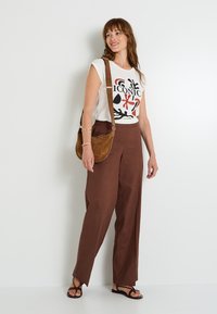 Camiseta gráfica blanca con el texto "ICÓNICO", pantalones de tiro alto marrones, bolsa de ante marrón, chanclas negras, de pie contra un fondo liso.